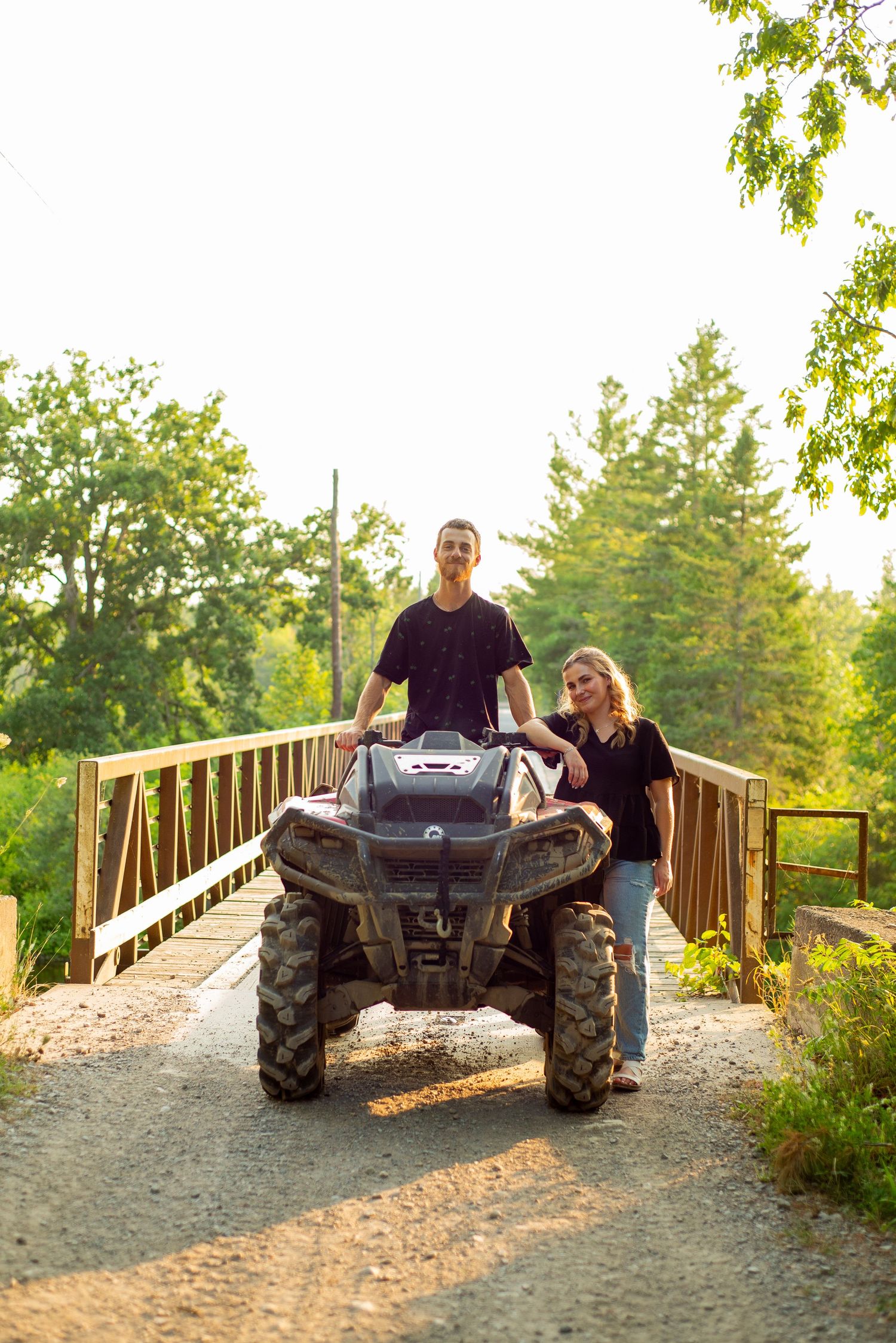 A couple stands with an ATV on a gravel path near a wooden bridge surrounded by lush green trees.