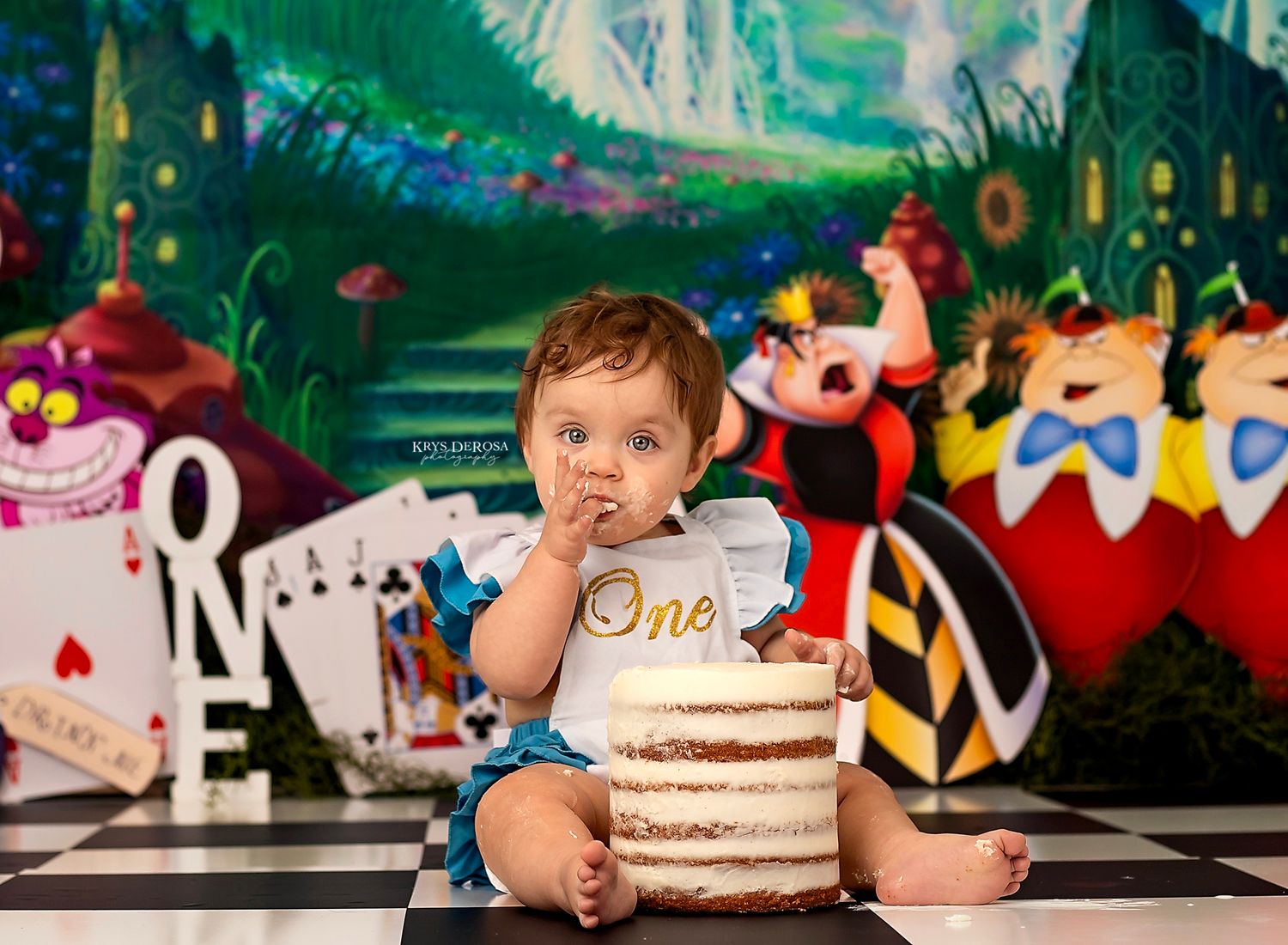 Baby girl in red outfit sits before a strawberry-themed cake smash backdrop with ONE letters for 1st birthday.