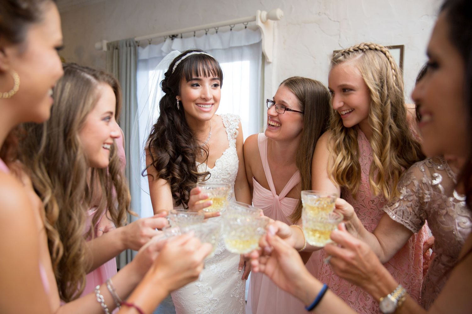 Bride in white gown toasts with bridesmaids in pink dresses, smiling and celebrating together indoors.