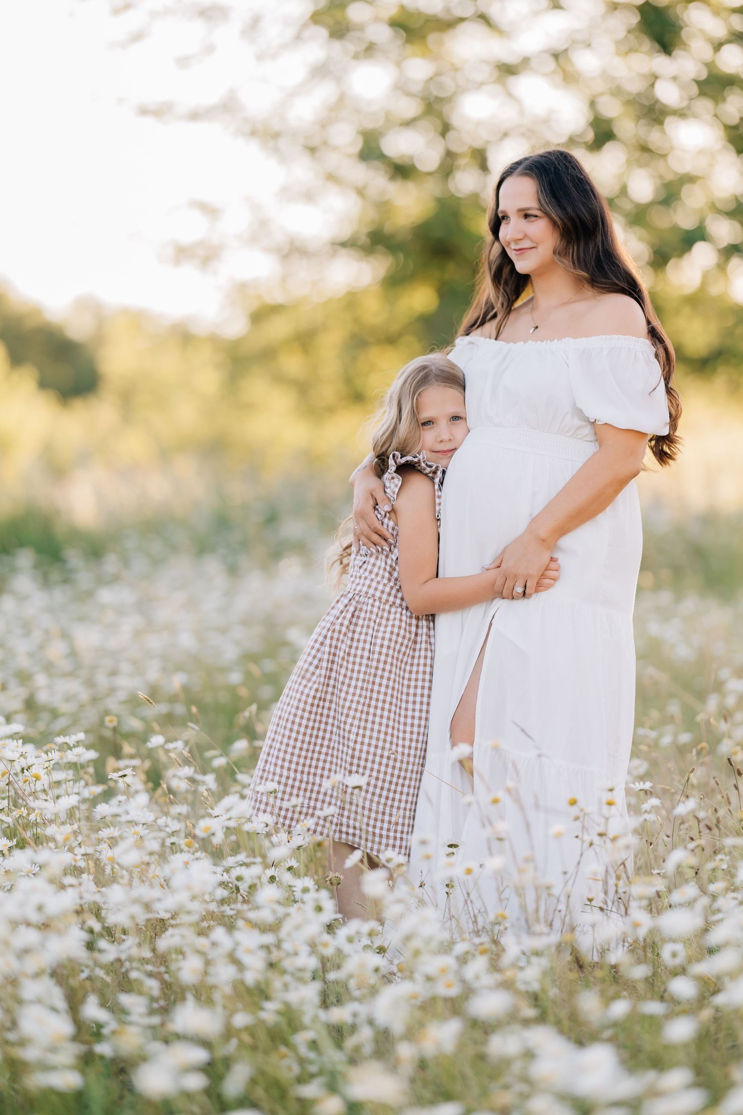 Pregnant woman in white dress with daughter in flower field at sunset.