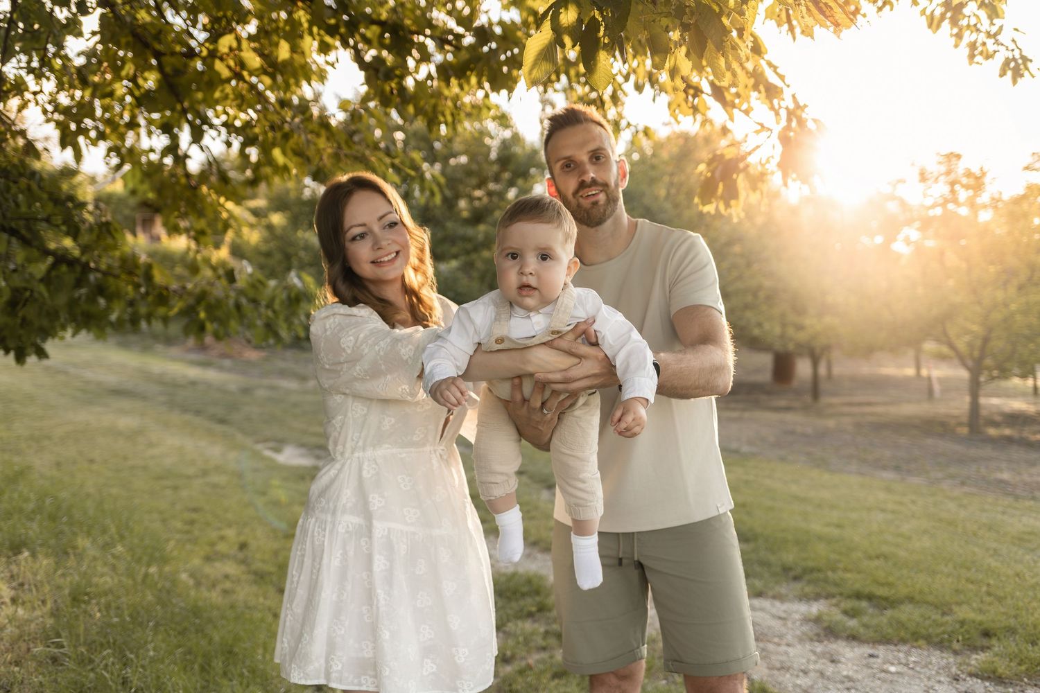 Young family with baby poses in golden sunset light among green trees in a park for a family portrait.