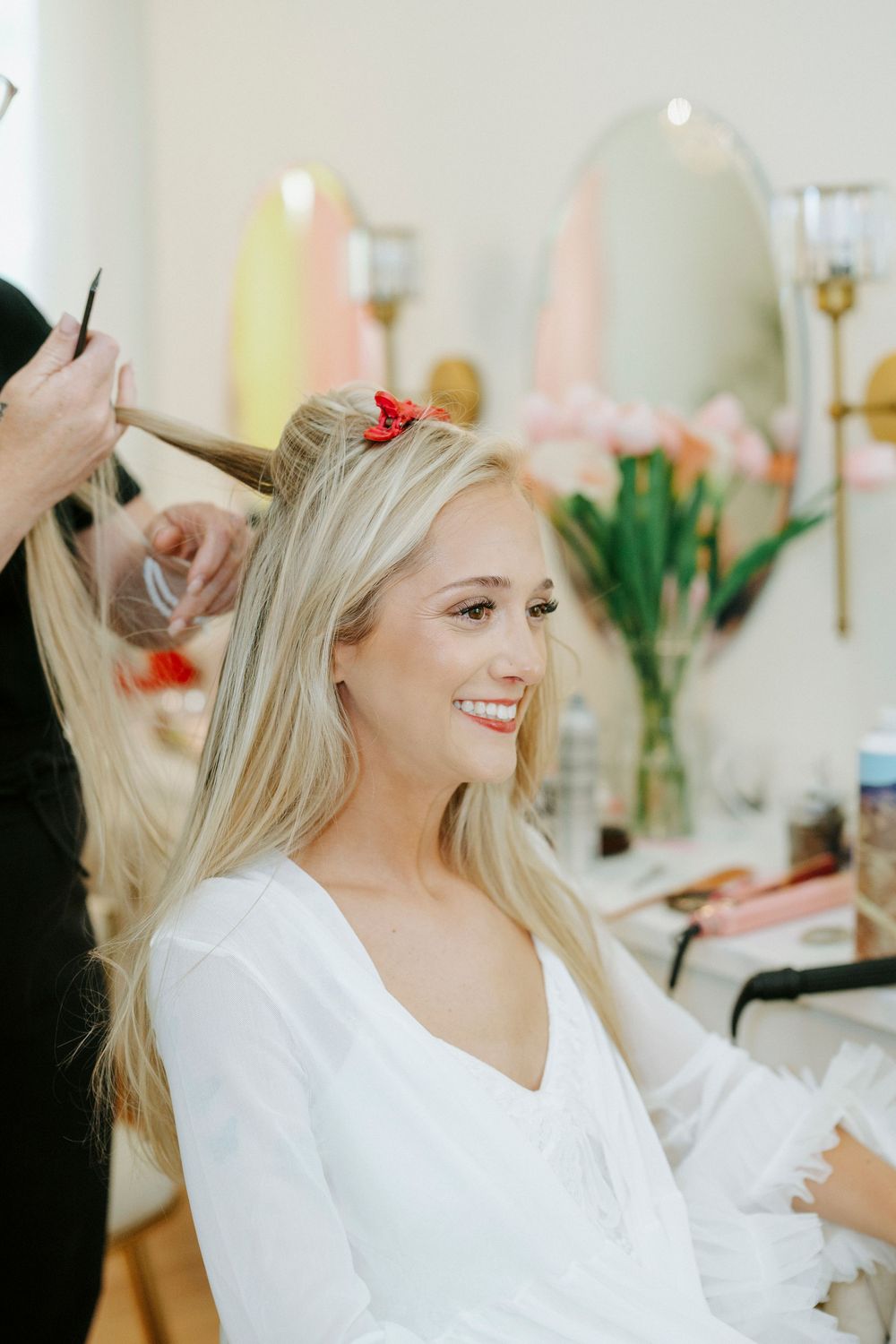 Smiling blonde bride getting hair styled in bright salon with floral decor on wedding day.