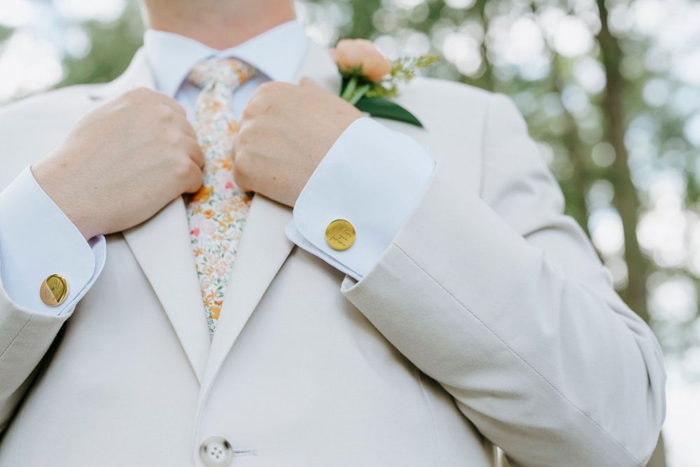Close-up of groom in beige suit adjusting floral tie with gold cufflinks and boutonniere.
