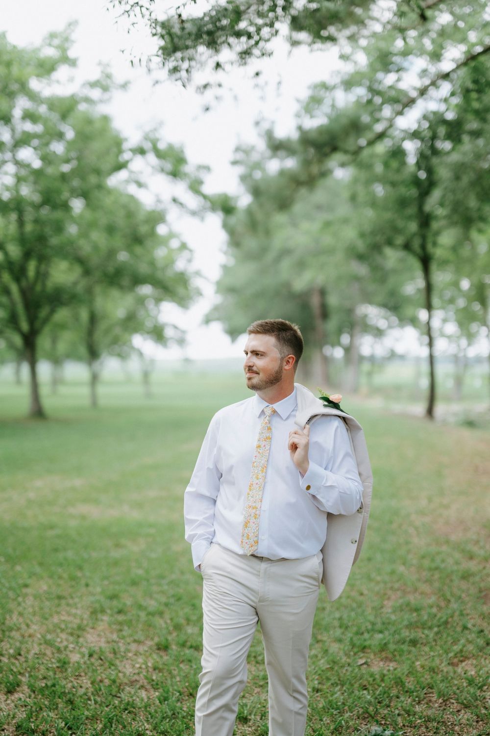 Groom in light gray suit holding jacket over shoulder in lush green park setting.