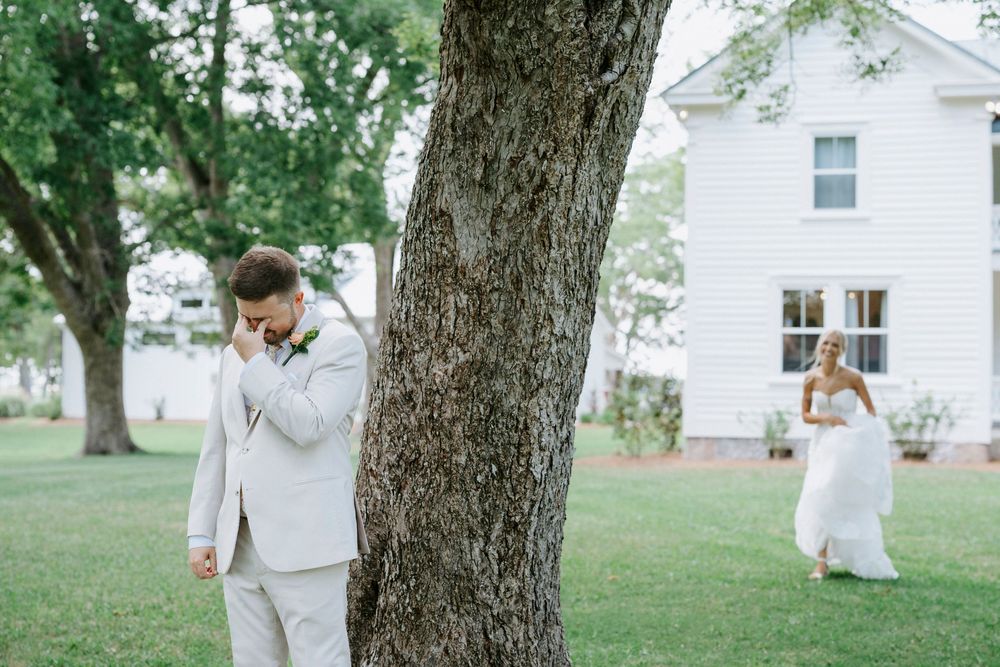 Groom wiping tears beside a tree during an emotional first look with bride approaching in white gown.