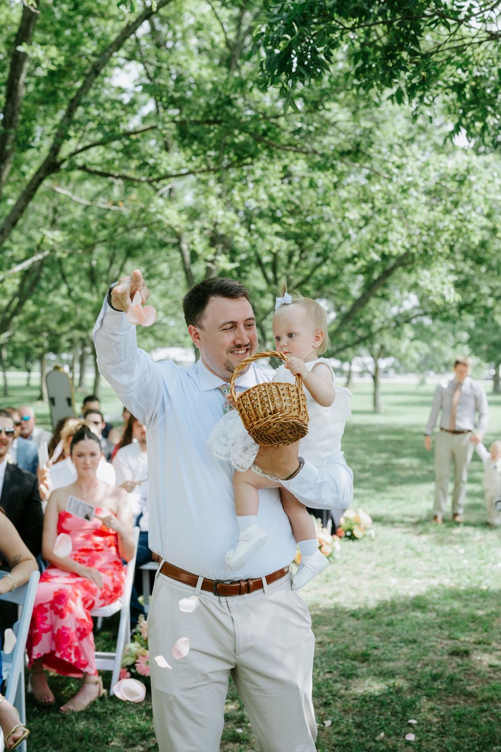 Father carries baby flower girl down outdoor wedding aisle while tossing petals at garden ceremony.