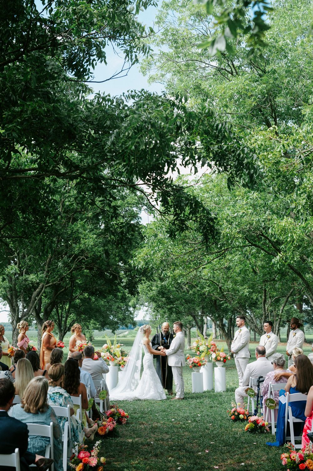 Outdoor wedding ceremony under lush green trees with colorful floral arrangements and seated guests.