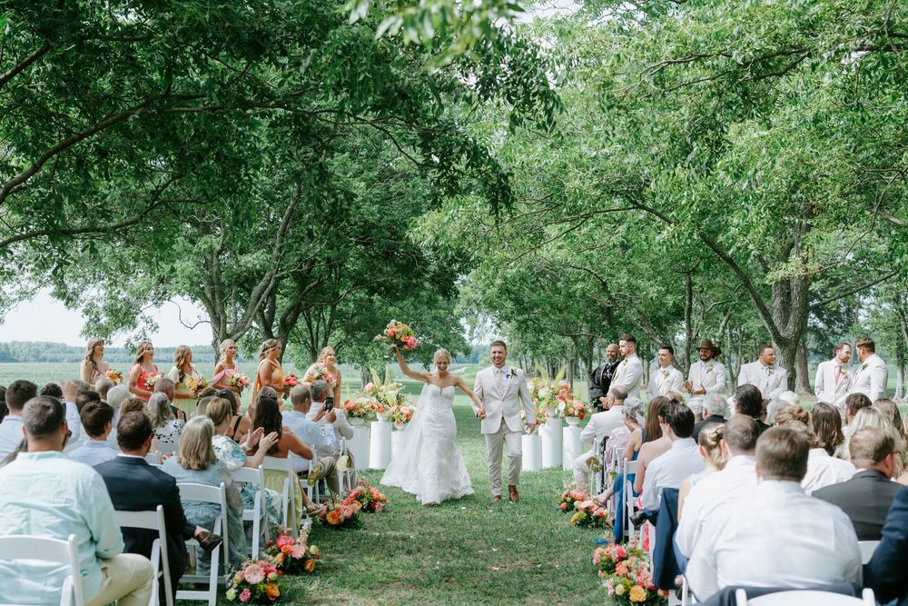 Outdoor wedding ceremony under lush green trees with guests seated and couple at the altar.