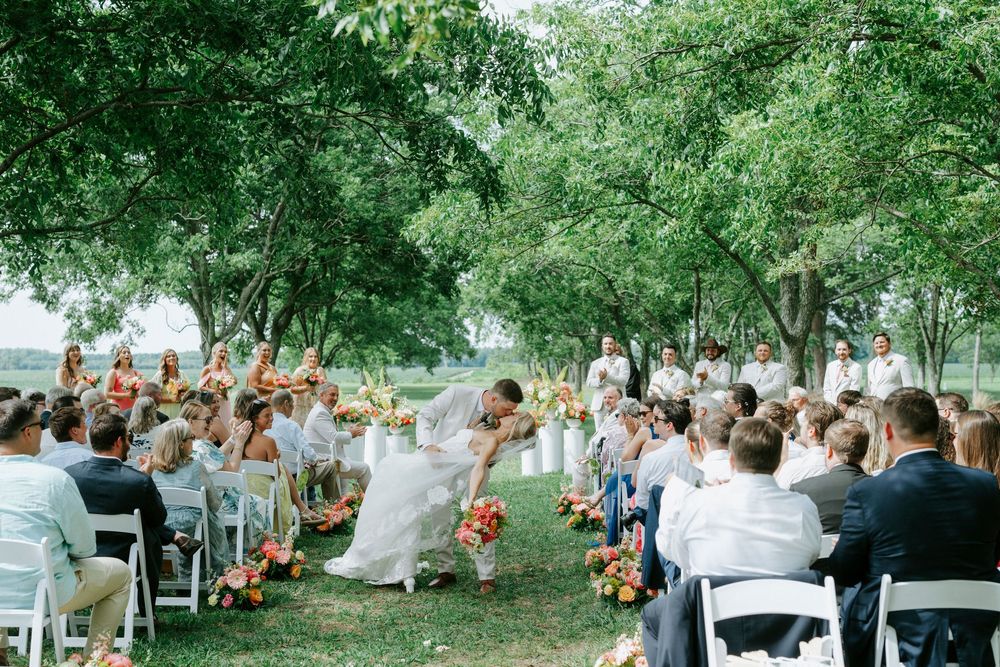 Bride and groom share first kiss at outdoor wedding ceremony under lush green trees.