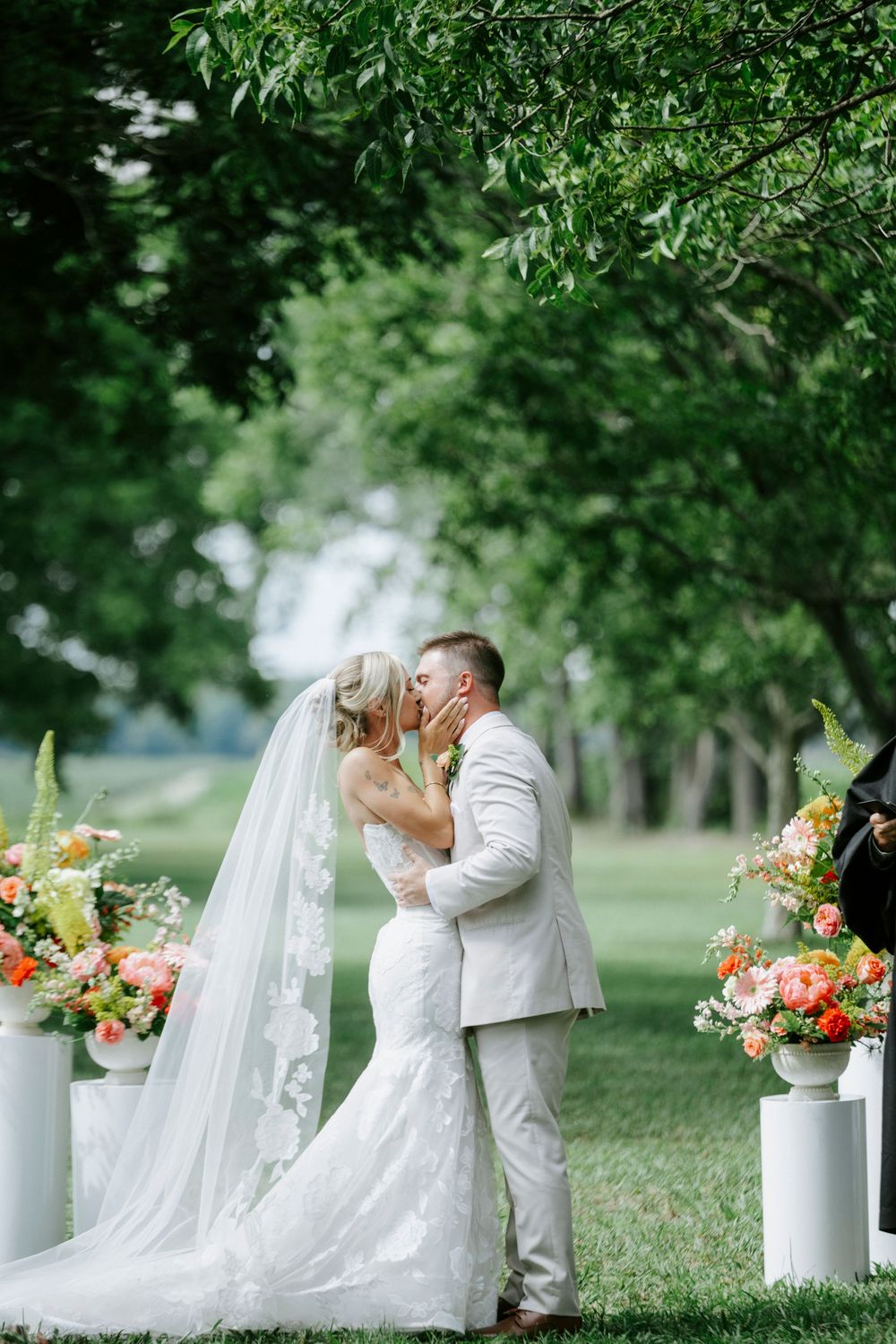 Bride and groom share first kiss at outdoor wedding ceremony flanked by colorful floral arrangements on white pedestals.