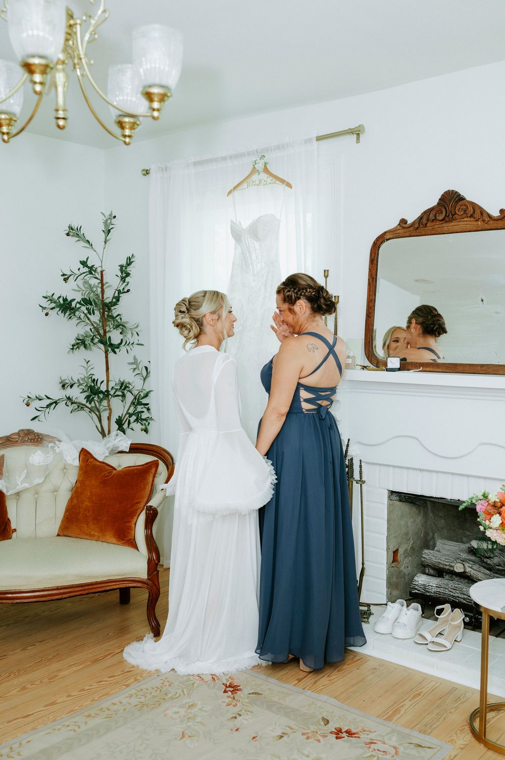Bride in white gown shares emotional moment with bridesmaid in blue dress inside elegant white bridal suite.