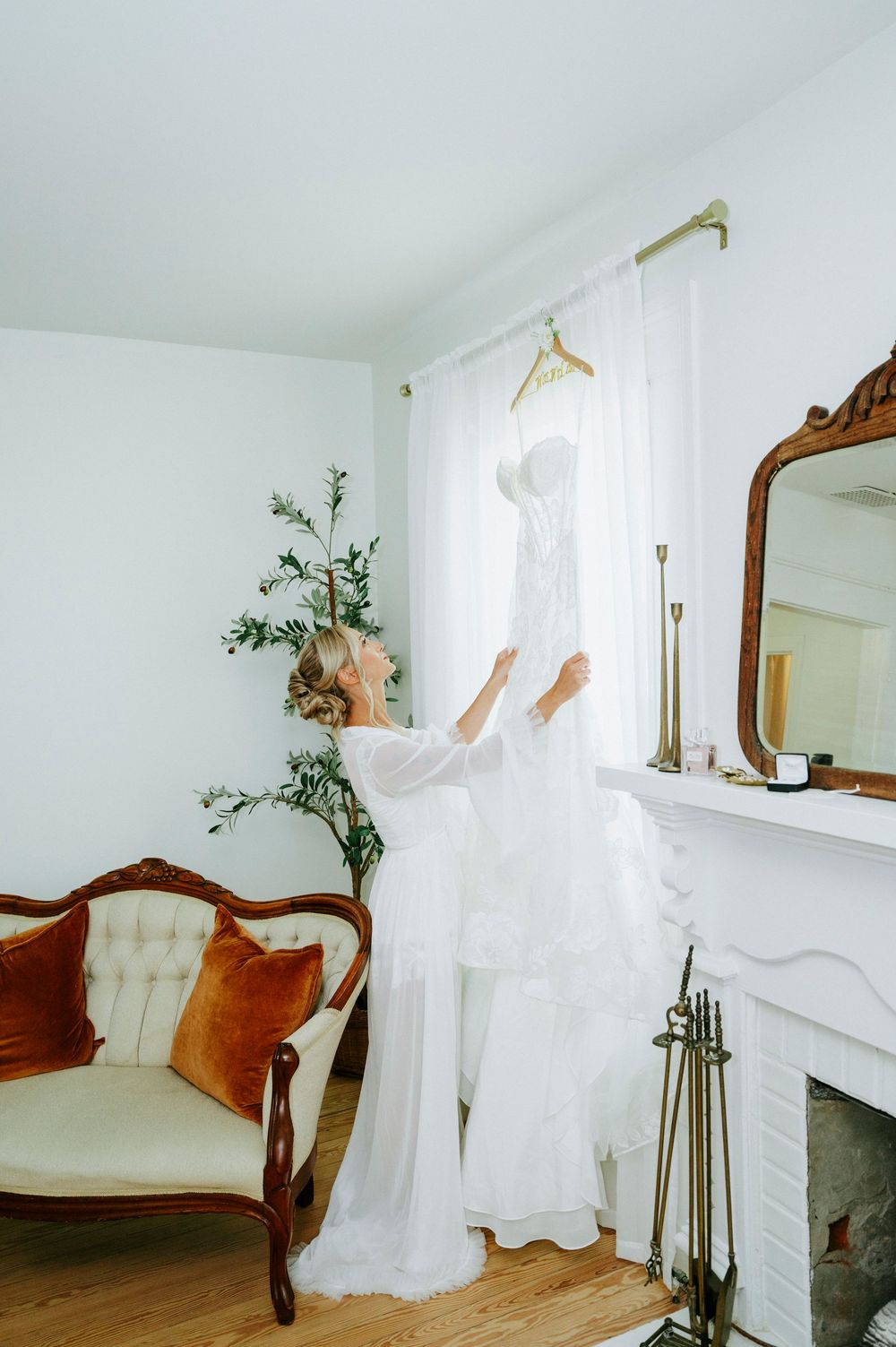 Bride in white robe reaching for her wedding dress hanging by a sunlit window in an elegant room.