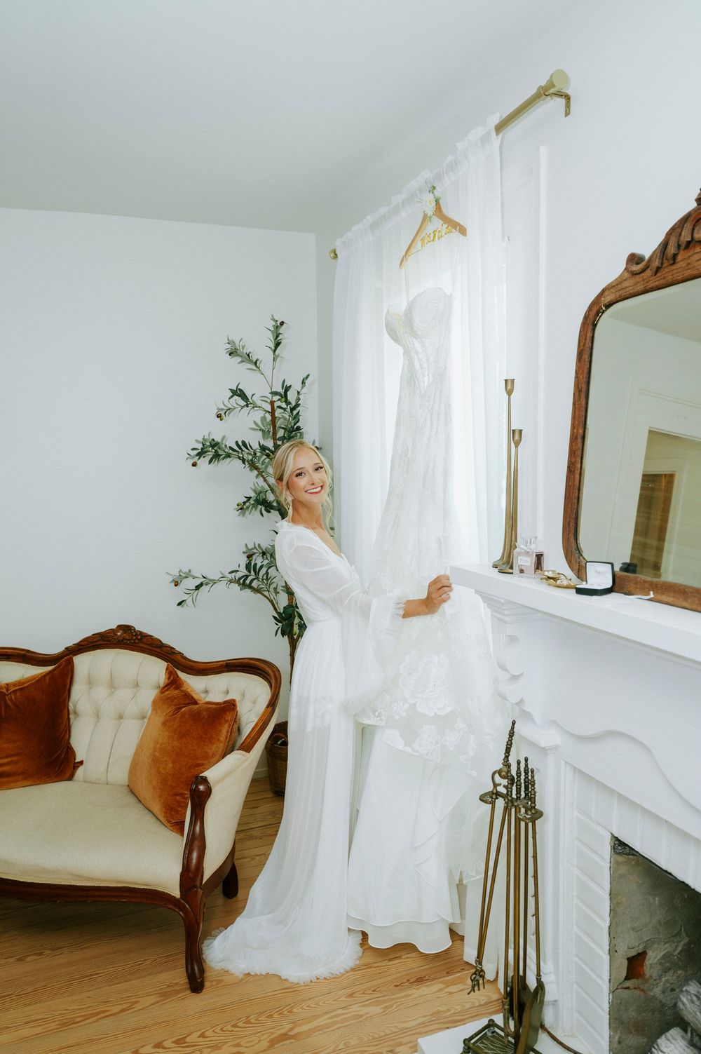 Smiling bride in white robe admires her wedding dress hanging by a fireplace in an elegant room.