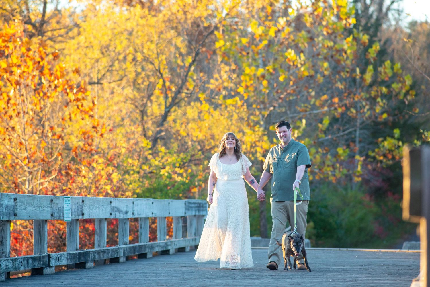 Couple in wedding attire walking hand-in-hand on a bridge with vibrant autumn foliage and a dog nearby.