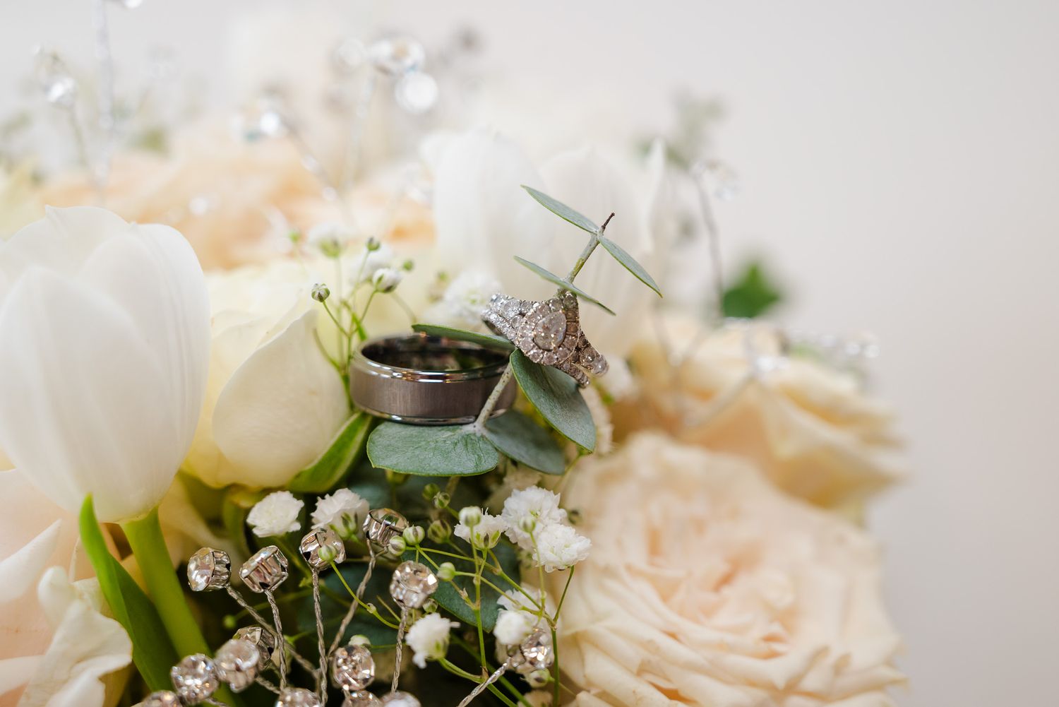 Wedding rings resting on a bouquet of white and blush roses with baby's breath and greenery.