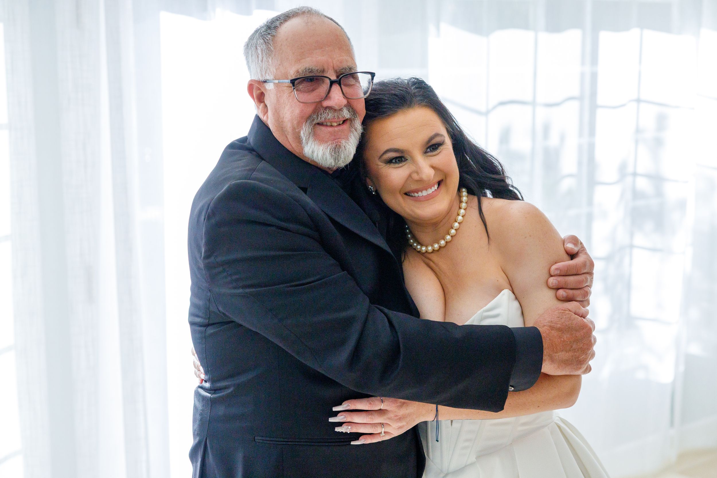 A bride in a white gown and pearl necklace hugs an older man in a dark suit, smiling warmly near a bright window.
