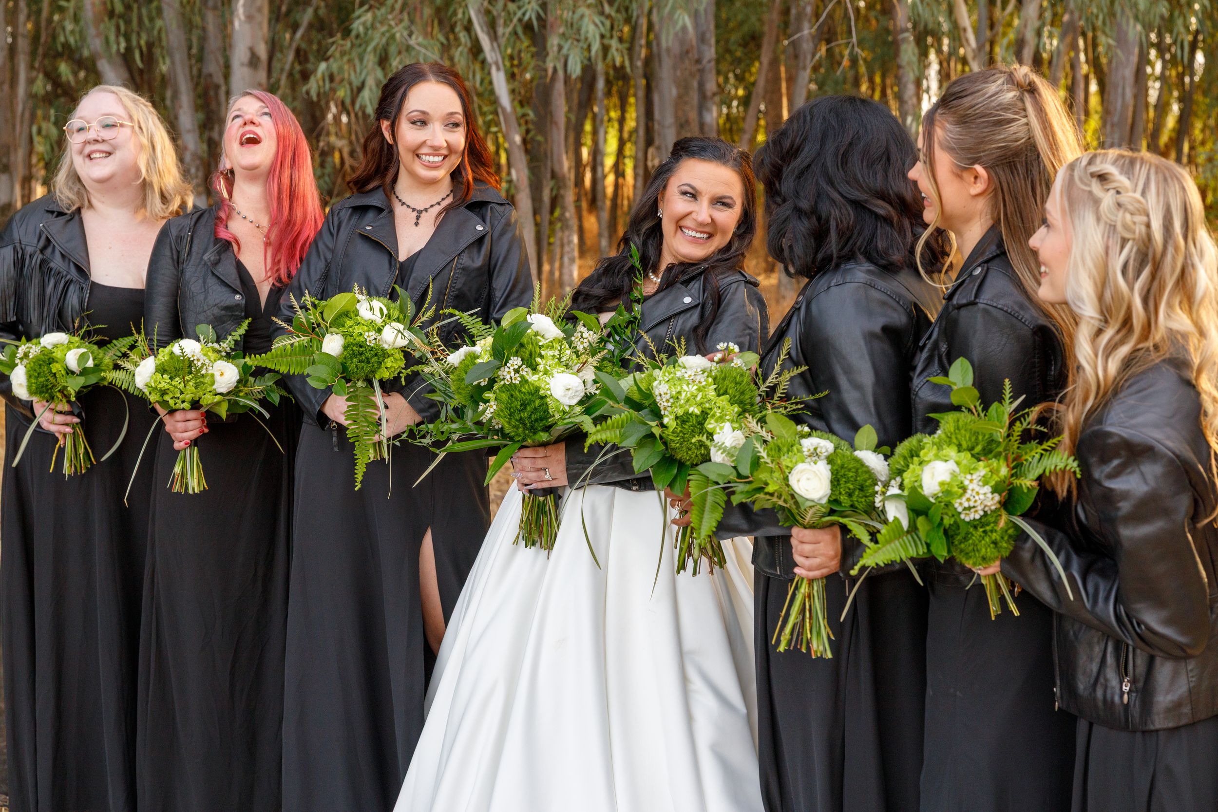 Bride in white gown with bridesmaids in black dresses and leather jackets holding white bouquets outdoors.