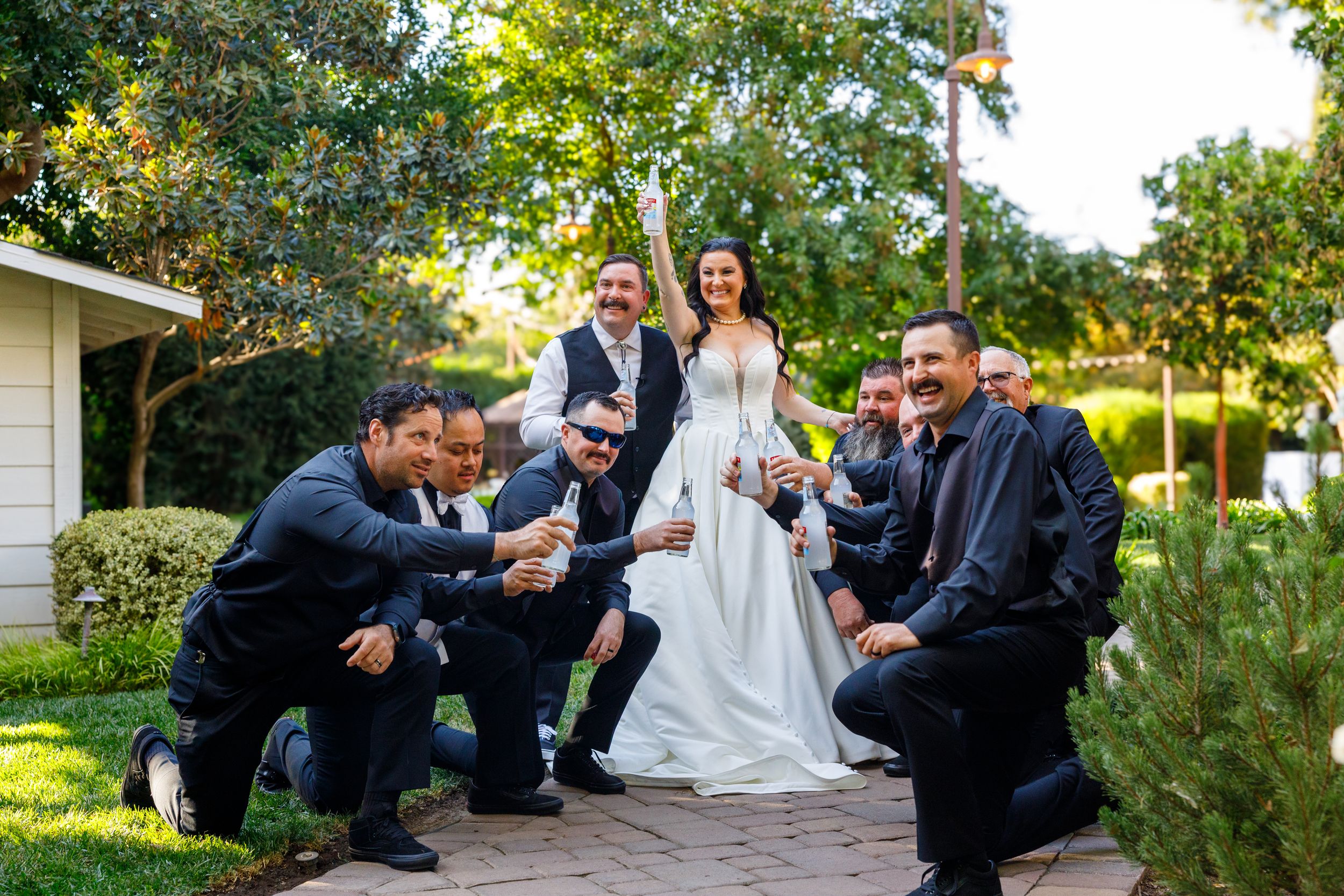 Bride and groom pose with groomsmen in black suits outdoors on a brick path surrounded by lush greenery.