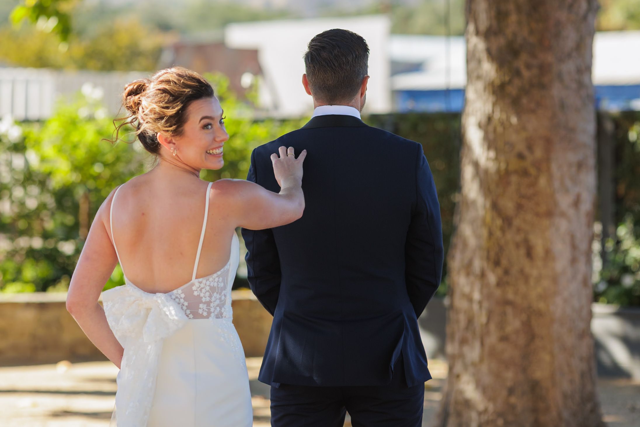 Bride in white gown touches groom's shoulder during outdoor first look wedding moment near a tree.