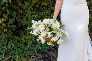 Bride in white gown holding elegant white floral bouquet with greenery backdrop at outdoor wedding.