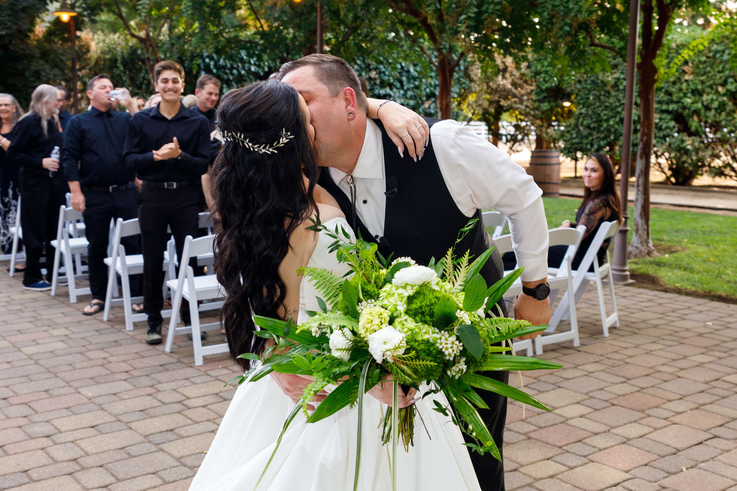Bride and groom share a kiss at outdoor wedding ceremony with white chairs and guests in background.