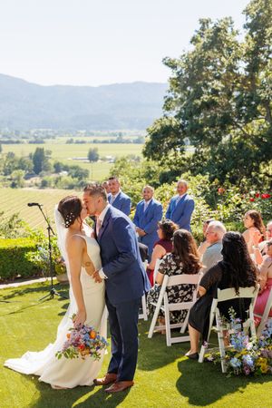 Bride and groom share first kiss at outdoor vineyard wedding ceremony with mountain views and seated guests.