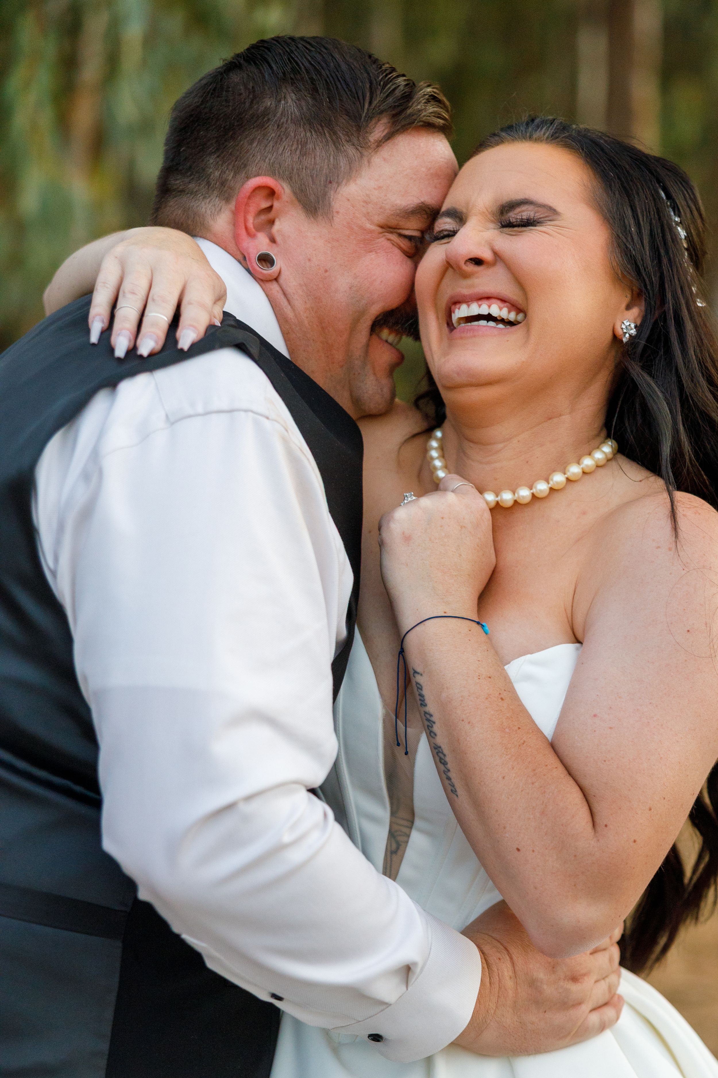 A joyful bride in a strapless gown and pearl necklace laughs as the groom nuzzles her cheek on their wedding day.