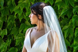Bride in white V-neck gown and sheer veil with pearl details, posing against lush green ivy backdrop.