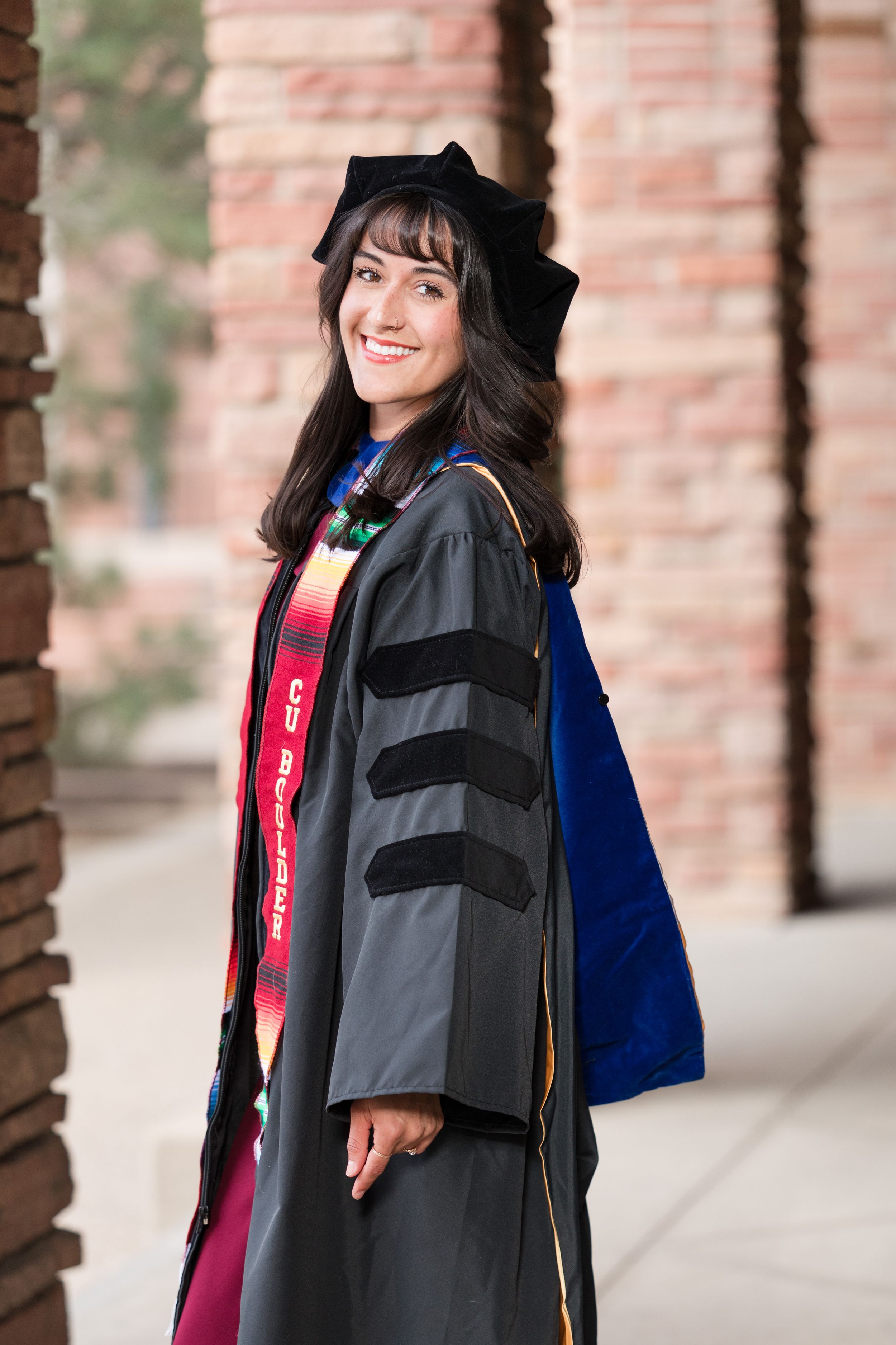 Smiling graduate in black doctoral gown, cap, and colorful regalia poses outdoors near brick columns.