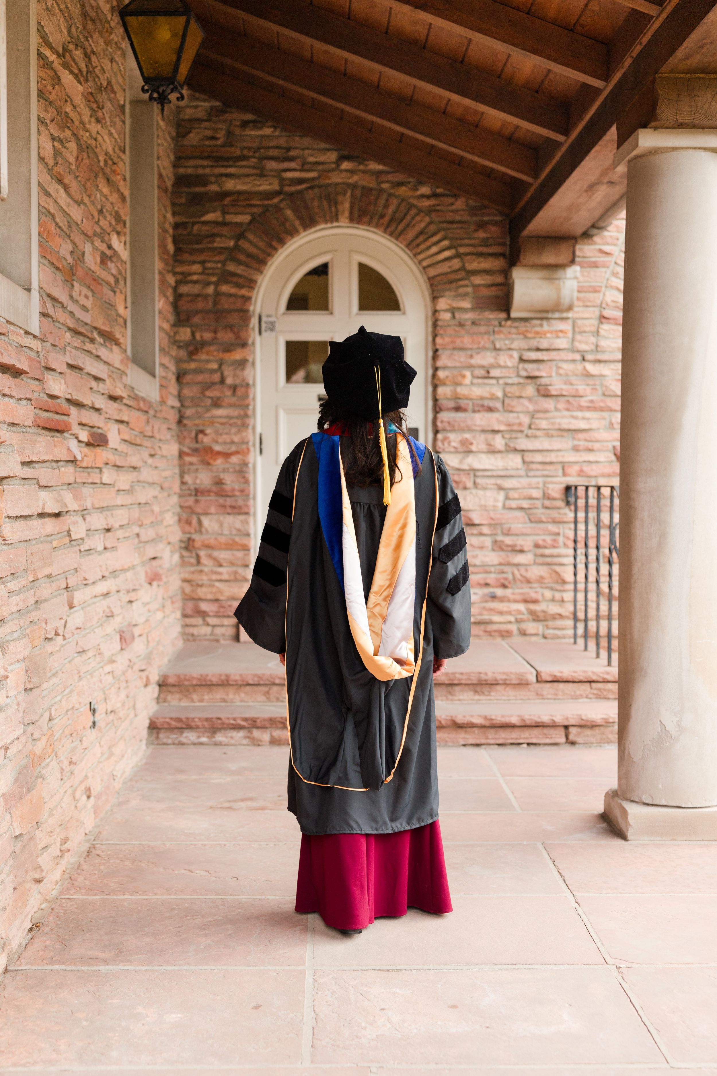 PhD graduate in black doctoral gown with colorful hood walks toward brick building entrance on campus.