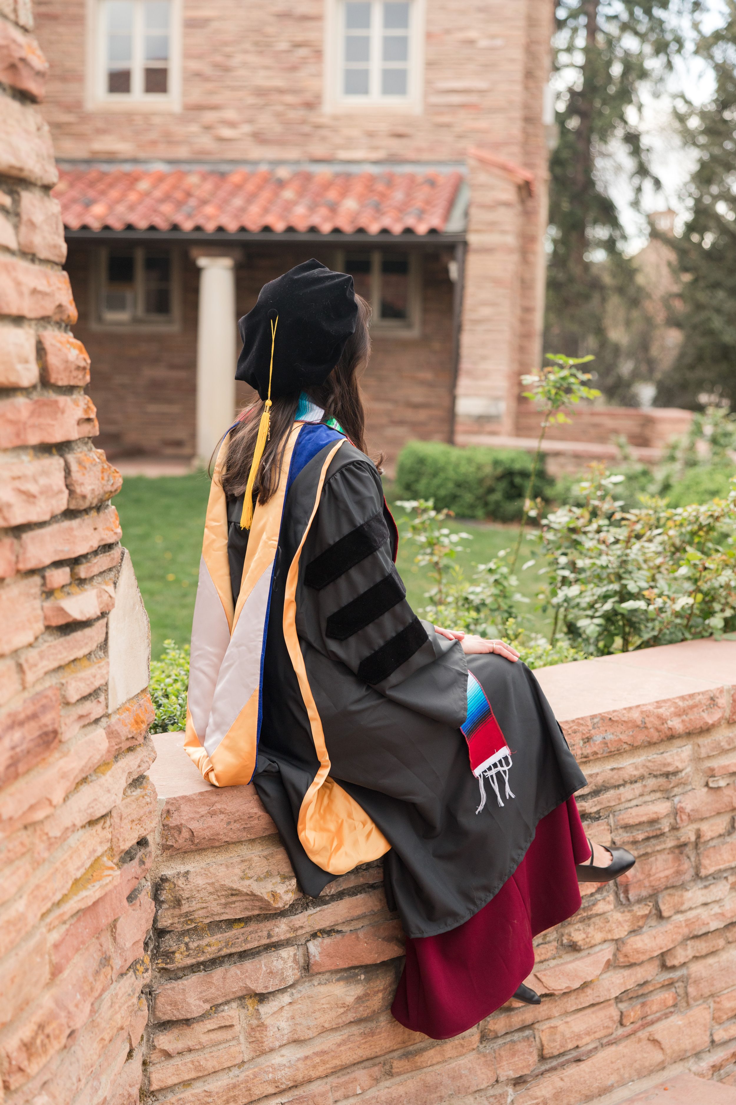 Graduate in black doctoral gown and cap sits on brick wall at university campus, viewed from behind.