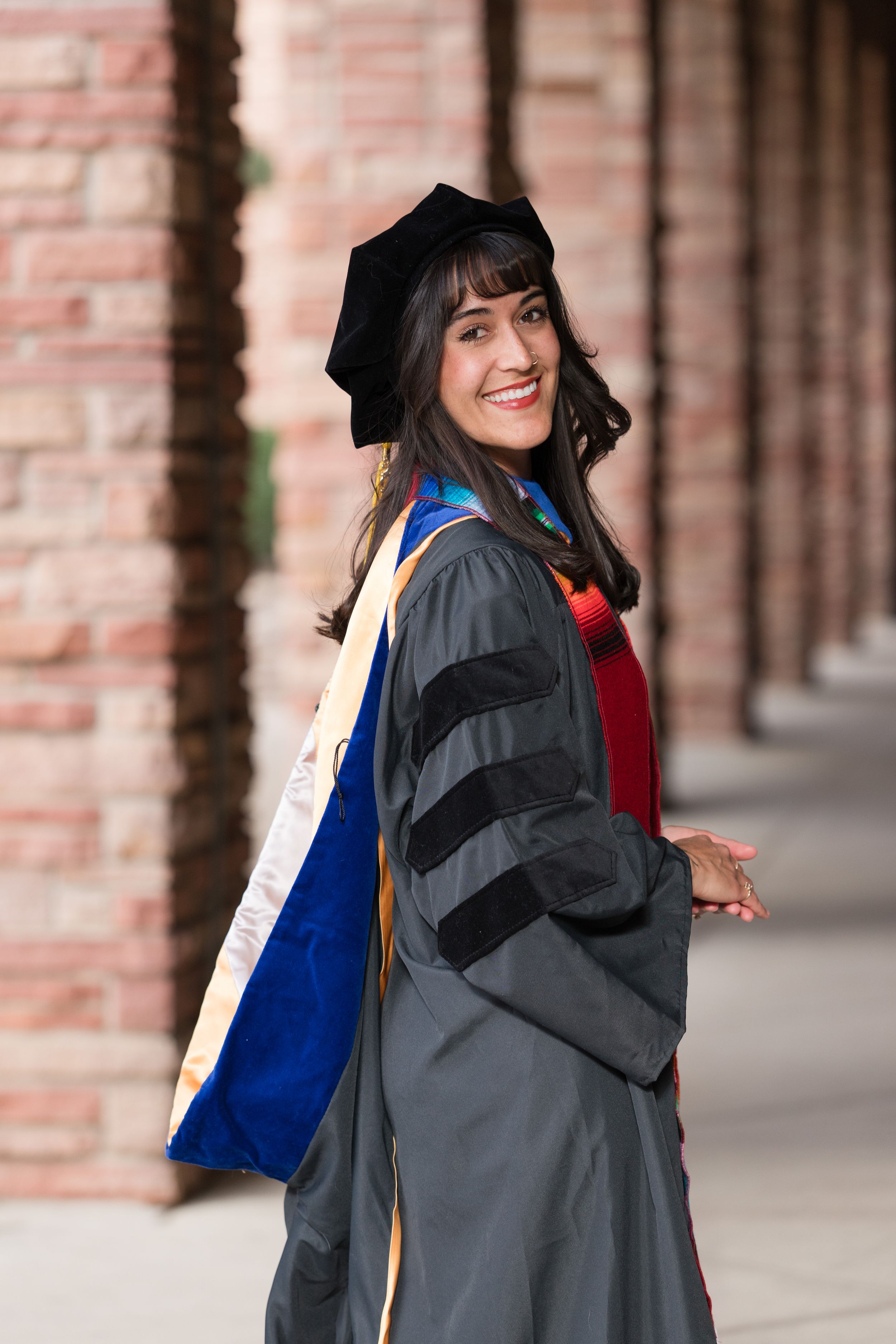 Smiling woman in doctoral graduation gown, cap, and colorful hood poses outdoors near brick columns.