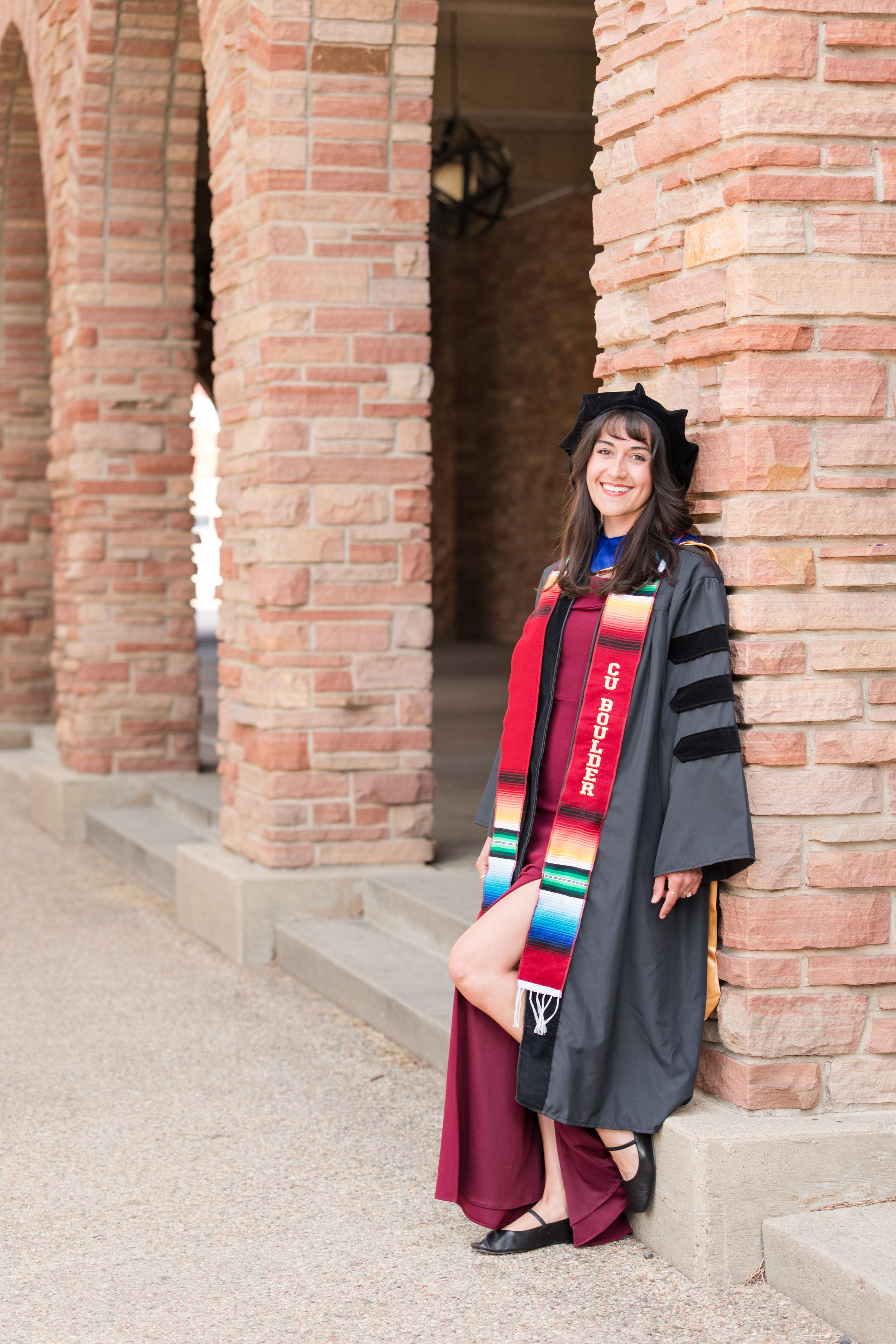 Woman in black doctoral graduation gown and colorful stole poses proudly by brick archway columns.