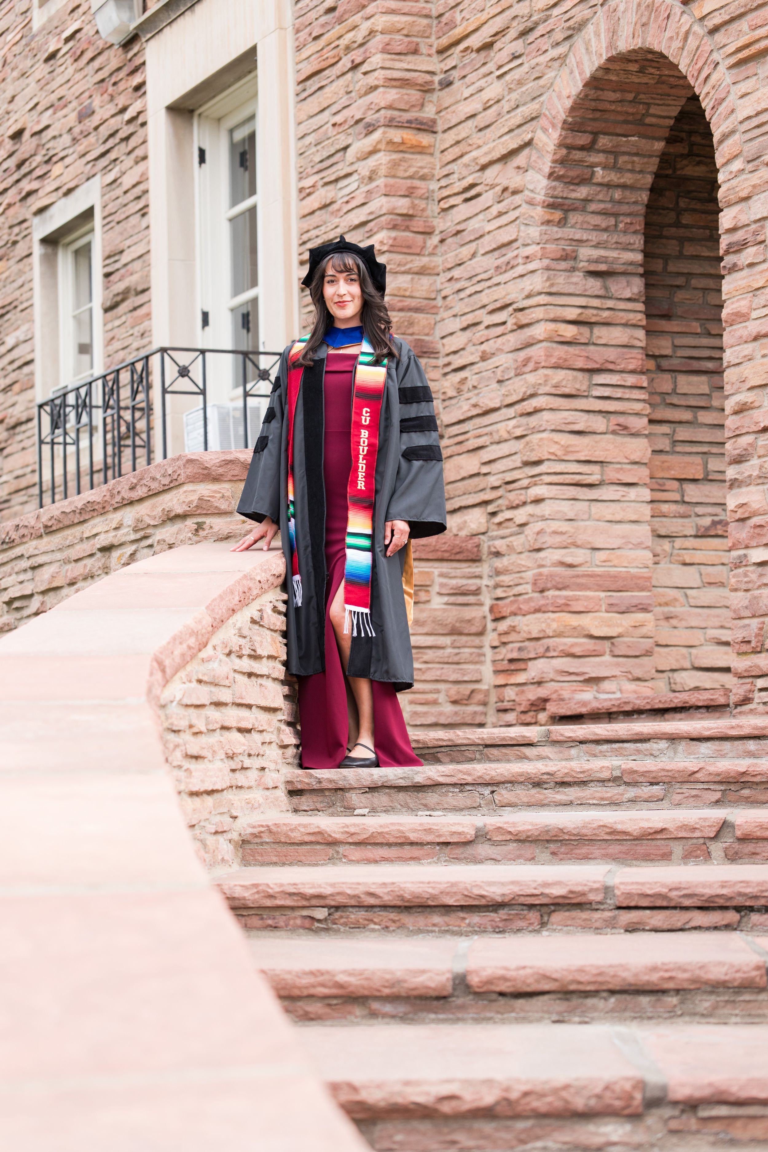 Graduate in cap and gown with colorful stole poses on stone steps outside a brick university building.