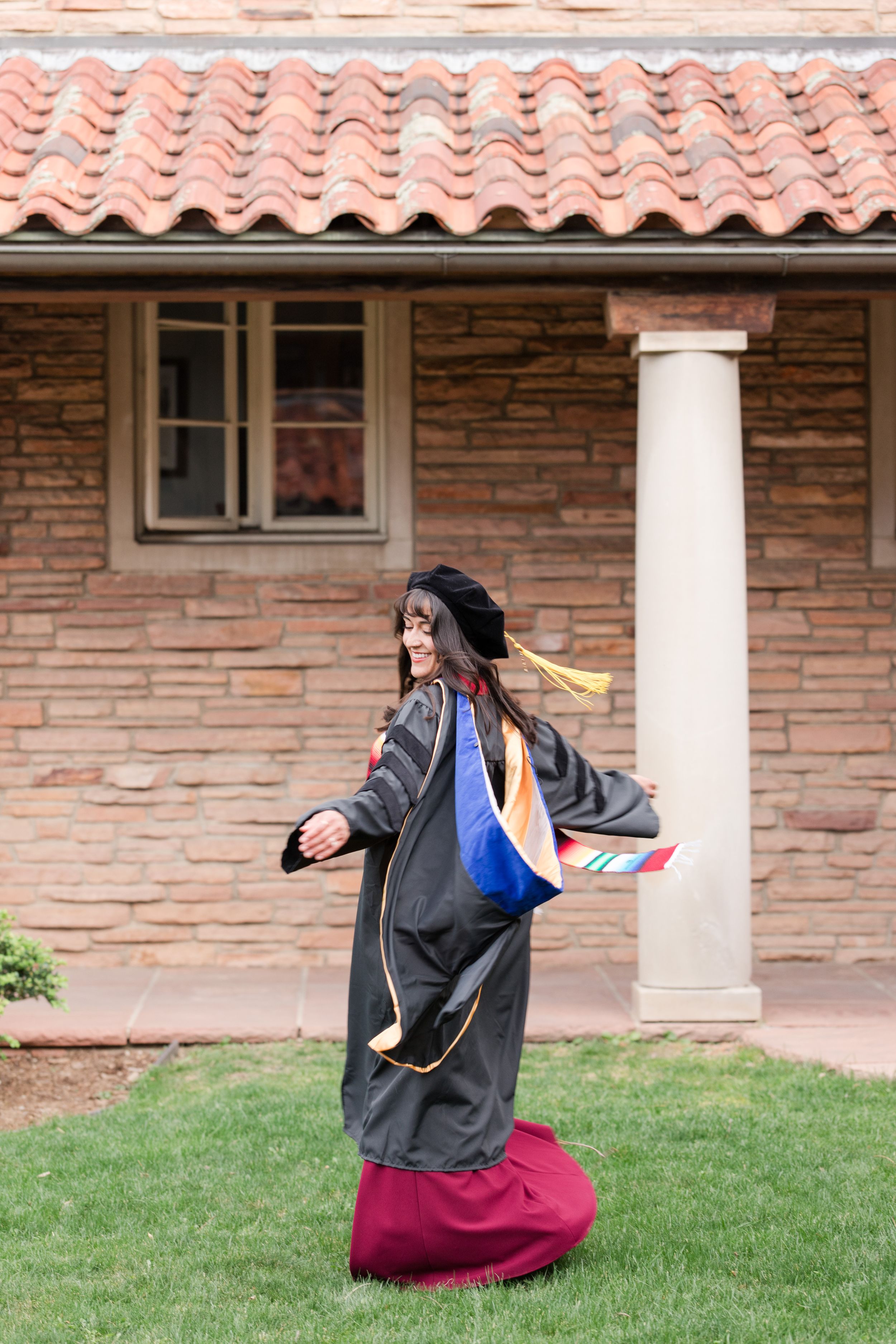 Woman in doctoral graduation regalia twirls joyfully on grass outside a brick building with terracotta roof.