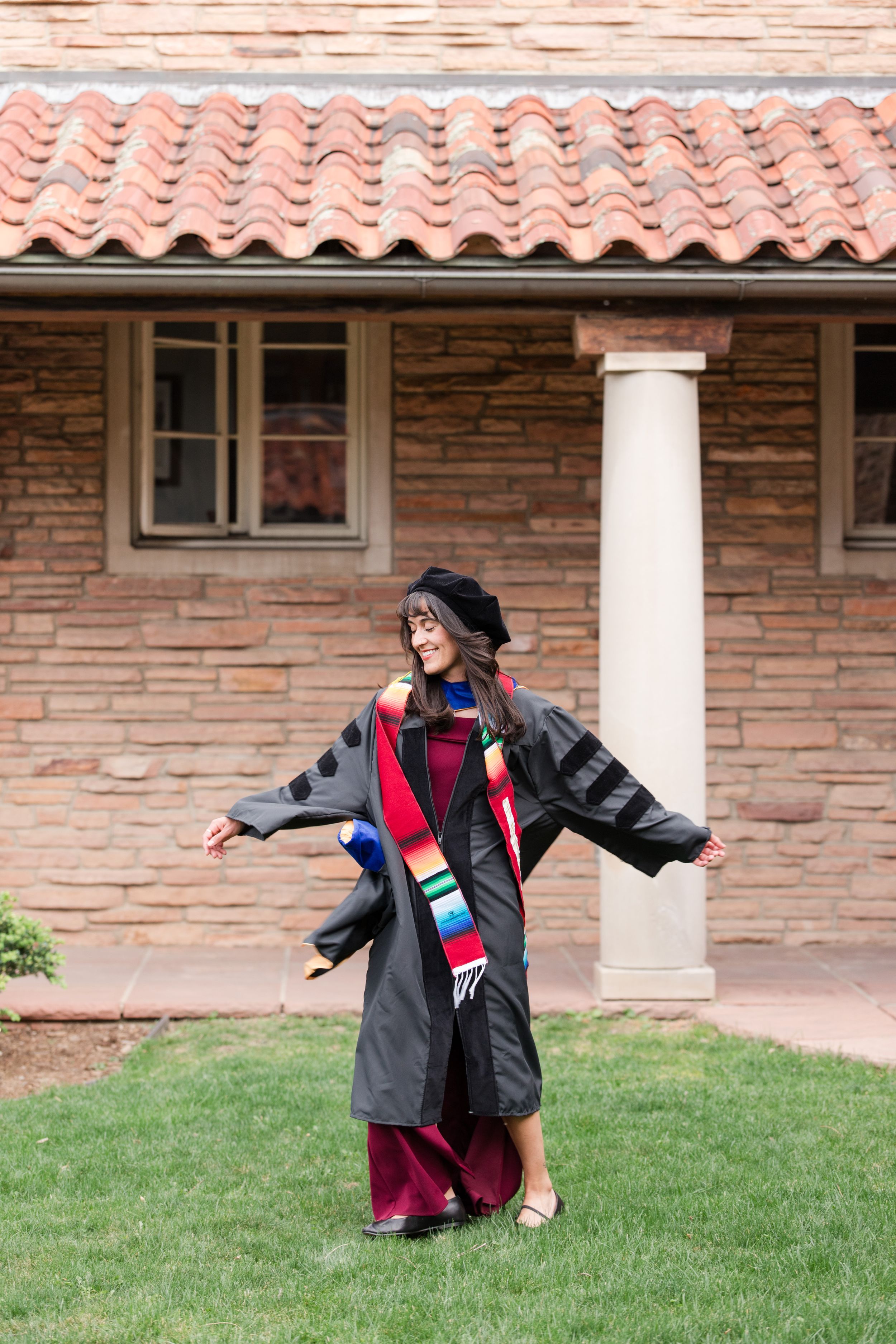 A joyful graduate poses outdoors in black doctoral regalia and colorful stole, arms outstretched, in front of a brick building.