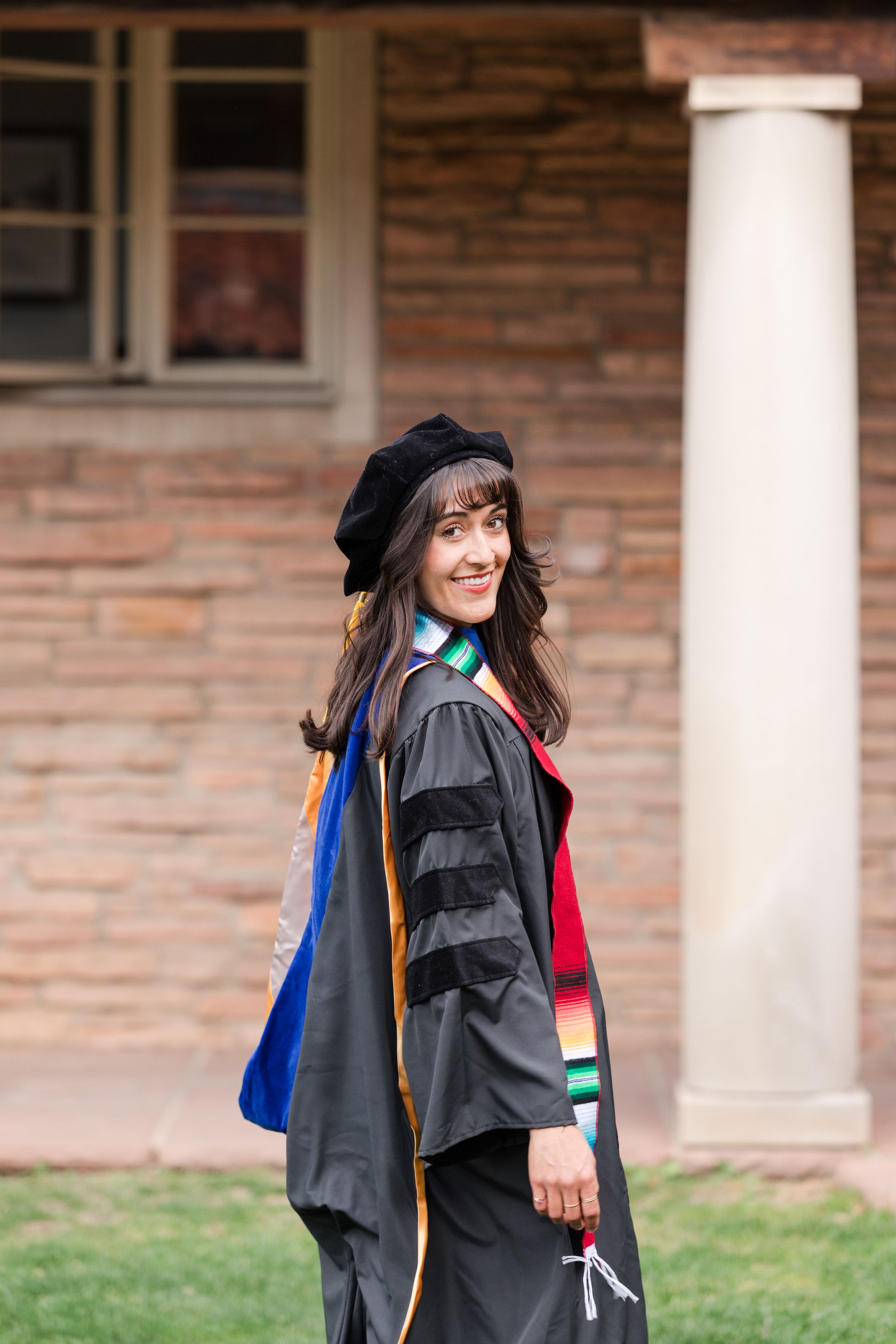Woman in doctoral graduation gown and cap smiling over shoulder outdoors near brick building.
