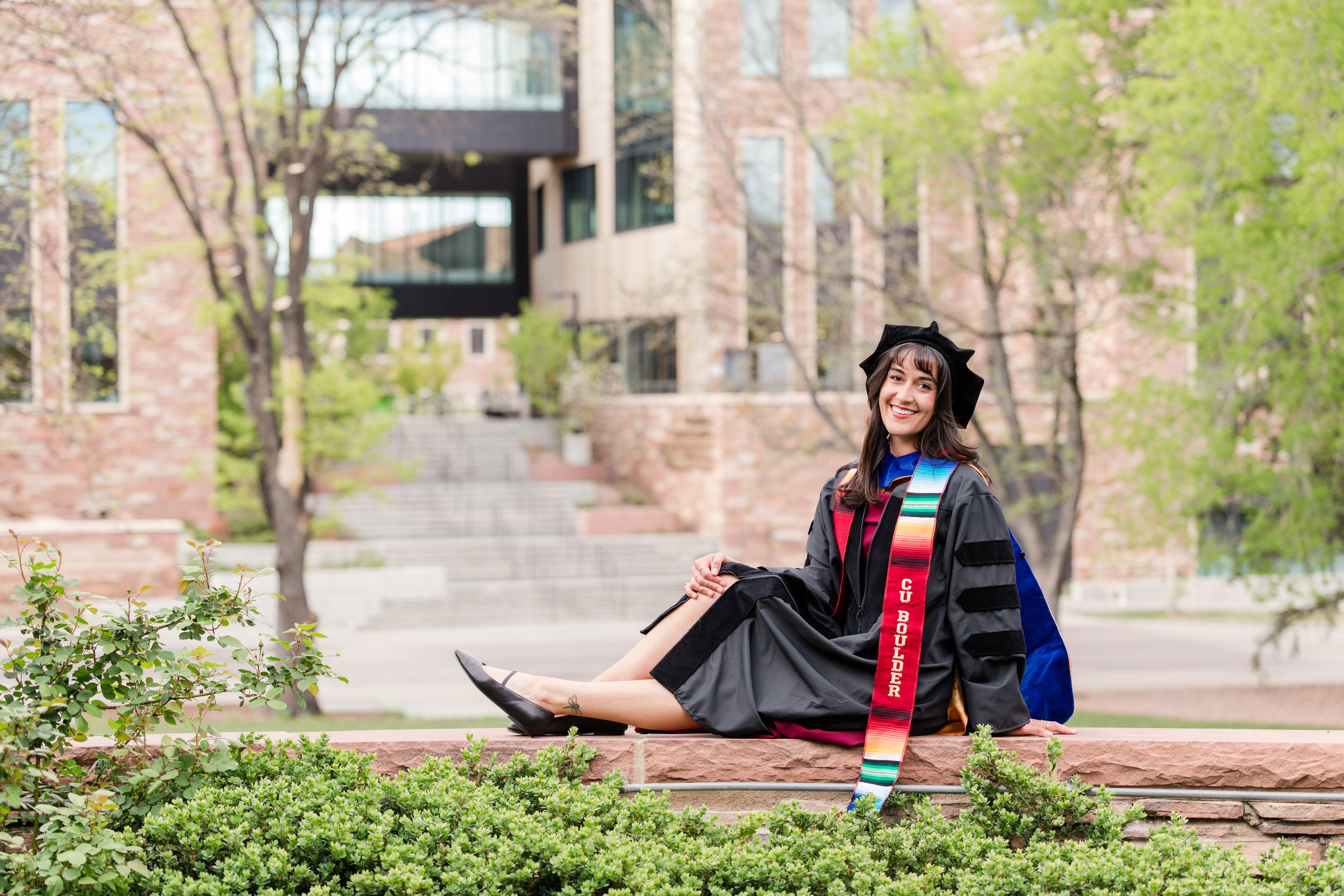 Woman in graduation cap and gown sitting outdoors on campus steps, smiling in doctoral regalia.