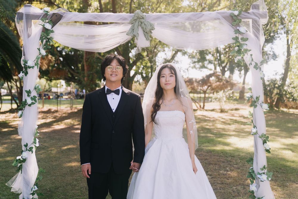 Smiling couple in wedding attire standing under floral arch outdoors, groom in black tuxedo, bride in white strapless gown.