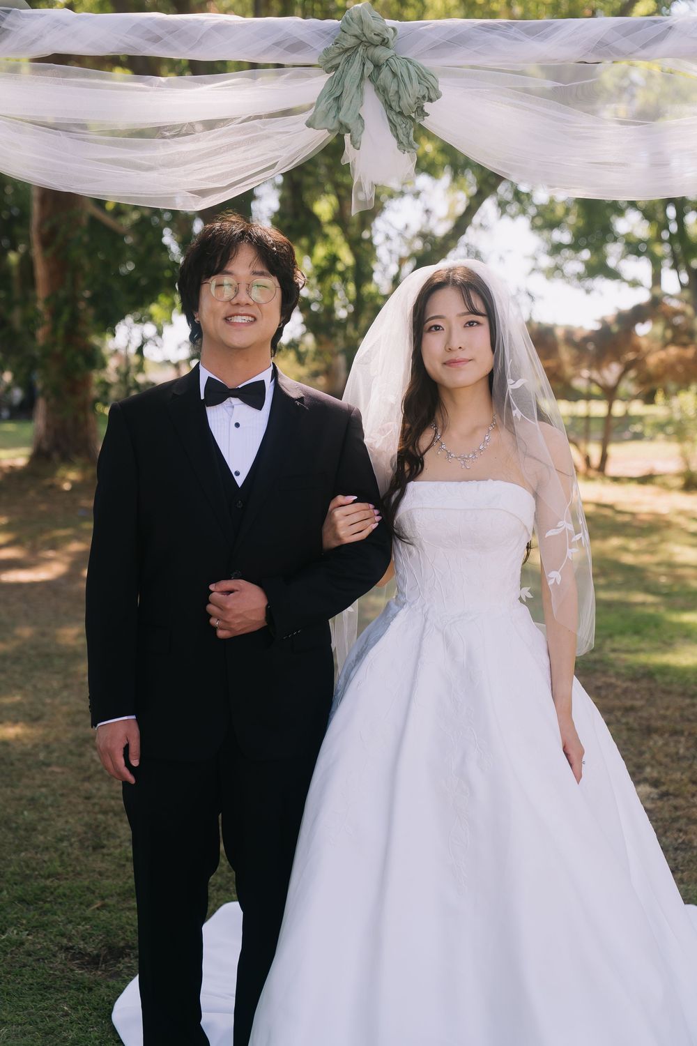 Couple posing at outdoor wedding ceremony, bride in white strapless gown with veil, groom in black tuxedo with bow tie.