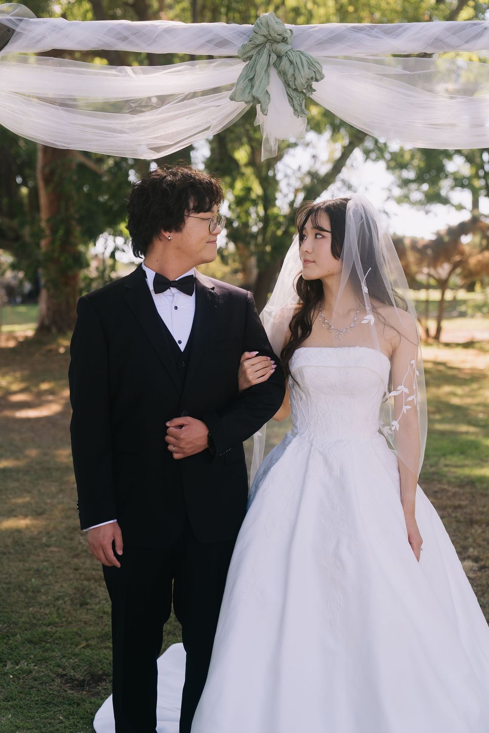 Wedding couple gazing at each other under draped arch outdoors, groom in tuxedo, bride in white strapless gown with veil.