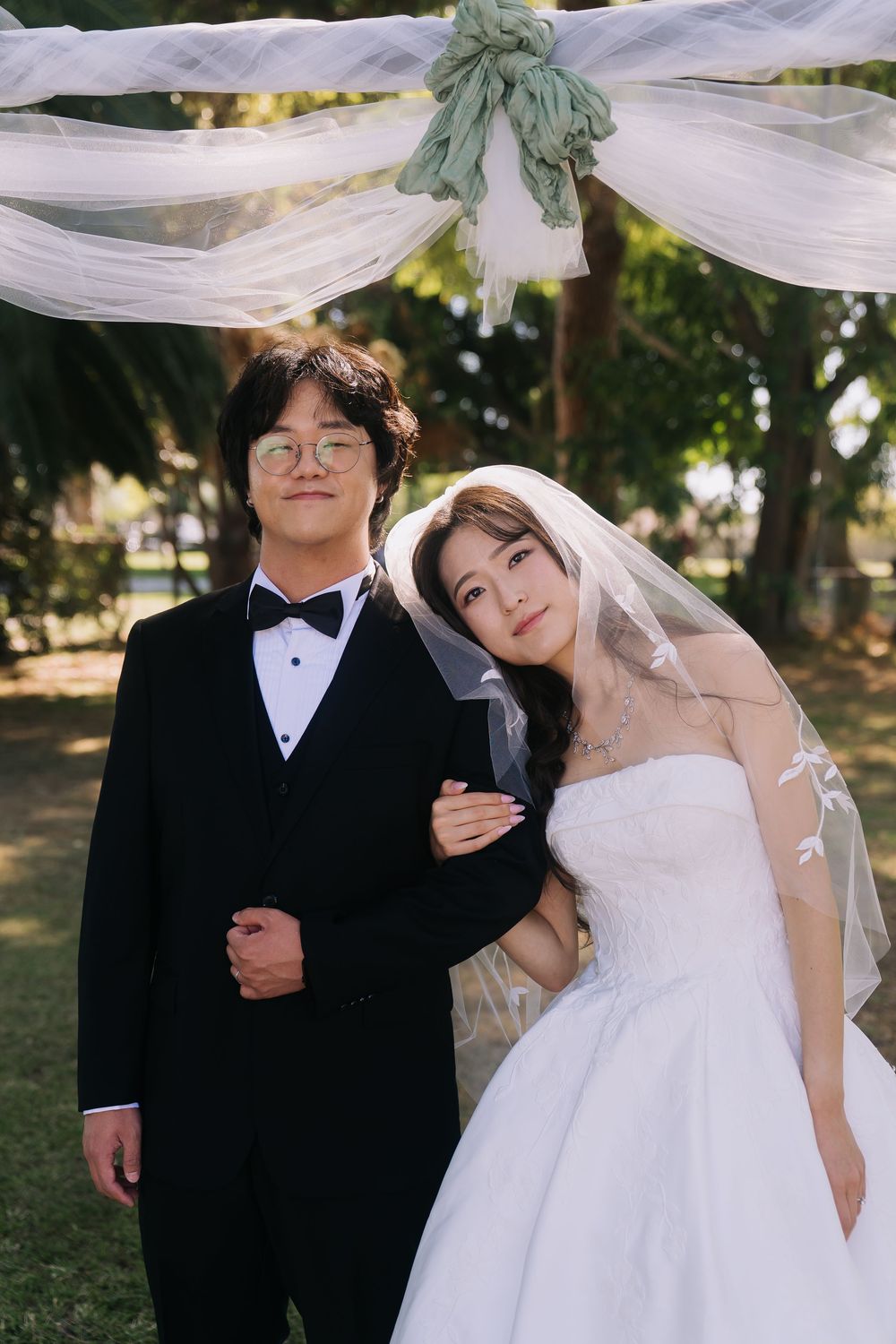 Couple posing in wedding attire under floral arch outdoors, groom in tuxedo and bride in white gown with veil.