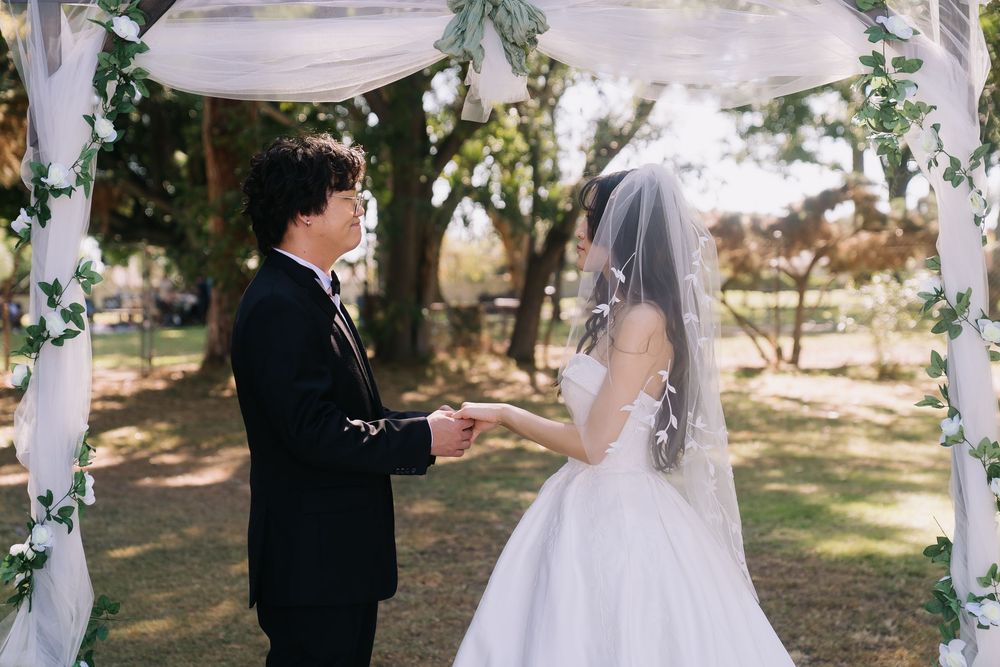 Bride and groom holding hands at outdoor wedding ceremony under decorated arch surrounded by trees.