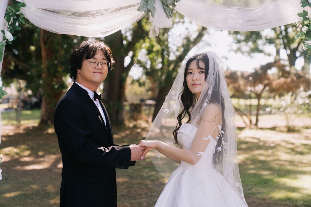 Couple exchanging vows at an outdoor wedding ceremony, bride in white gown with veil, groom in black tuxedo.
