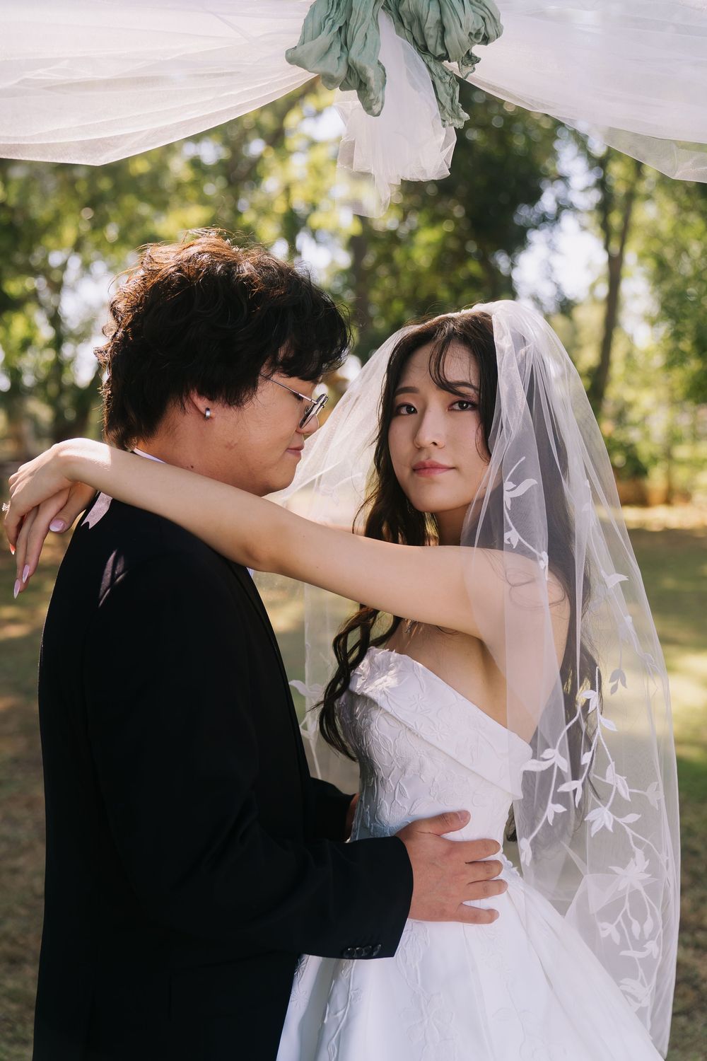 Asian bride in white gown and veil embraces groom in black suit outdoors at elegant garden wedding ceremony.