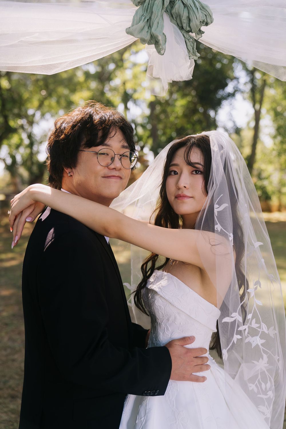 Asian couple in wedding attire pose outdoors, bride in white gown and veil embracing groom in black suit.