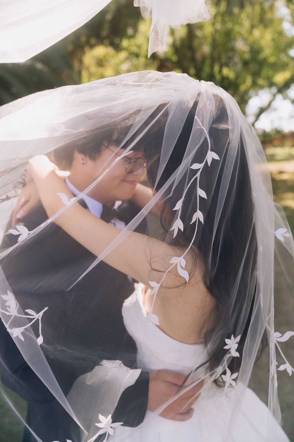 A bride and groom share a romantic moment under a floral-embroidered wedding veil outdoors.