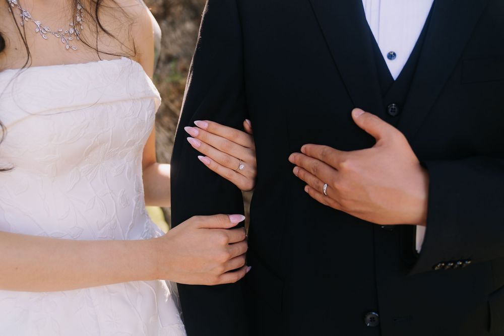 Bride and groom showing wedding rings, wearing white dress and black suit at outdoor ceremony.