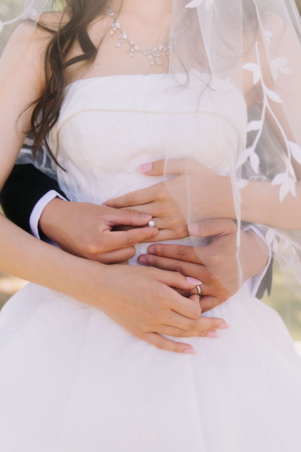 Bride and groom hands with wedding rings embracing at waist, bride in white dress with veil and diamond necklace.