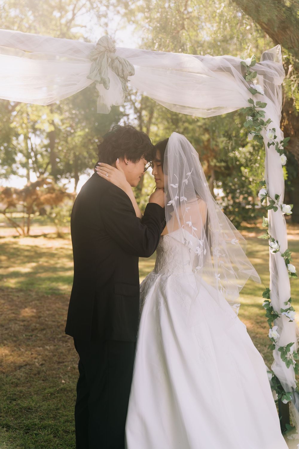 A bride and groom share an intimate moment under a floral arch at an outdoor wedding ceremony.